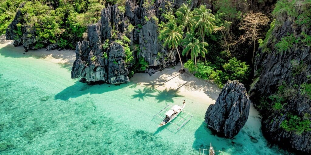 Cala escondida de El Nido desde el aire: paredes de piedra caliza con vegetación, palmeras sobre la arena blanca y una barca bangka fondeada en agua turquesa