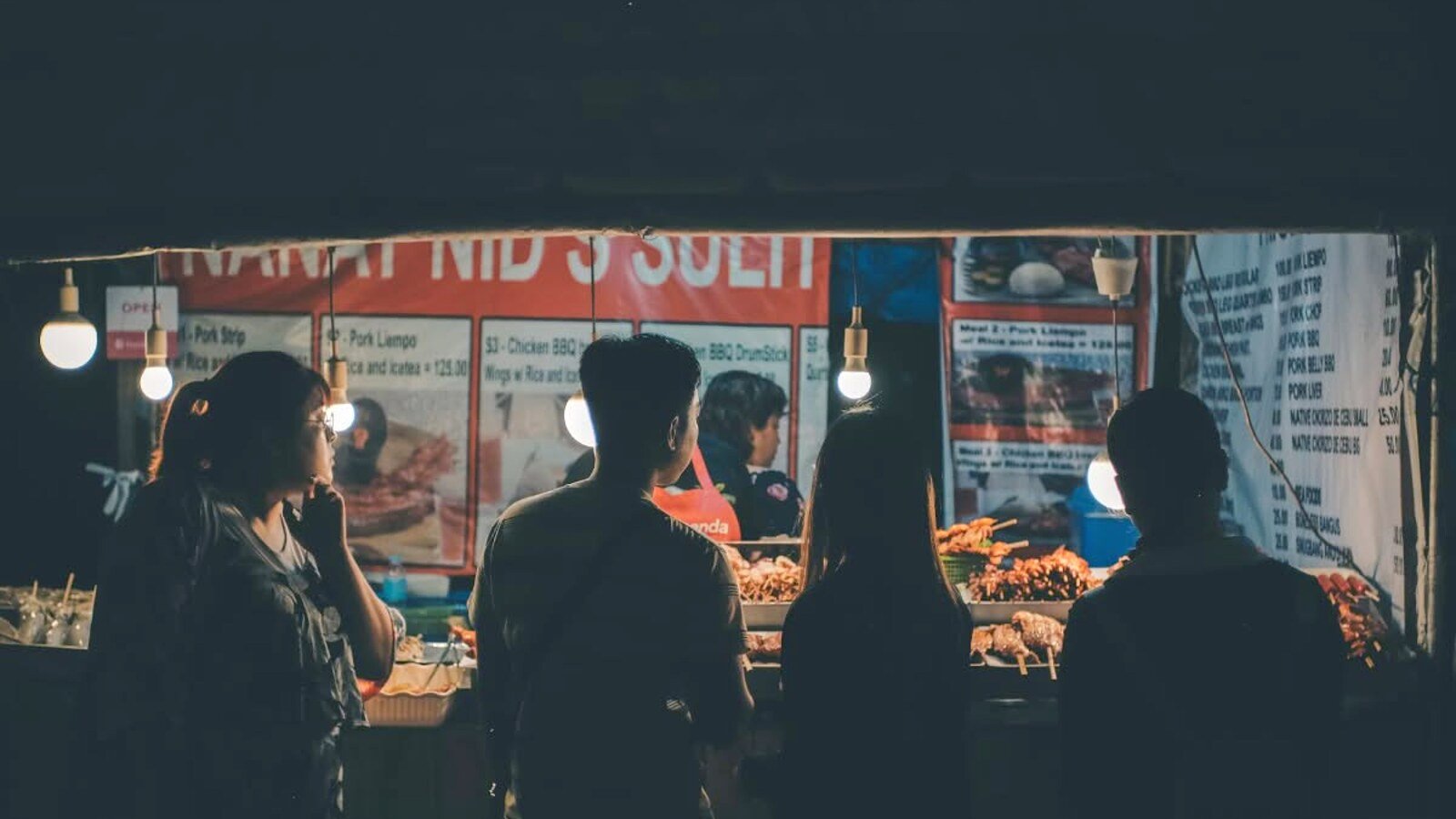 Puesto de comida callejera filipina iluminado de noche, con clientes eligiendo brochetas y platos a la brasa